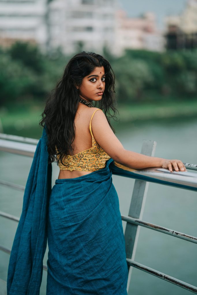 A woman in a traditional saree stands on a bridge, gazing back with a cityscape view.