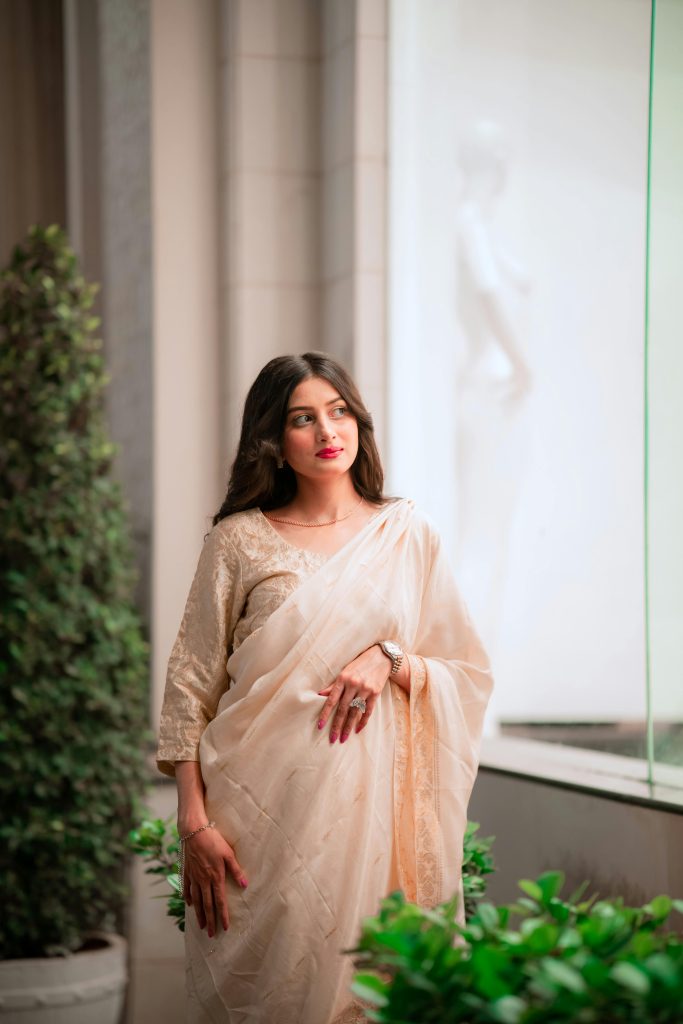 A woman wearing a traditional saree stands elegantly by a window, offering a blend of modern and classic style.
